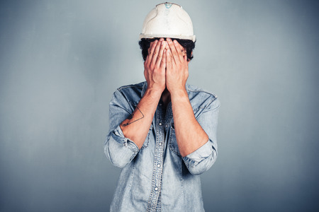 A blue collar worker wearing a hardhat is covering his face with his handsの写真素材