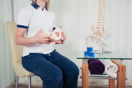 A physiotherapist is sitting in her office and is holding a model of the human pelvisの写真素材