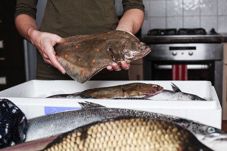 A young woman is in her kitchen and is holding a plaice with other types of fish in a box in front of herの写真素材