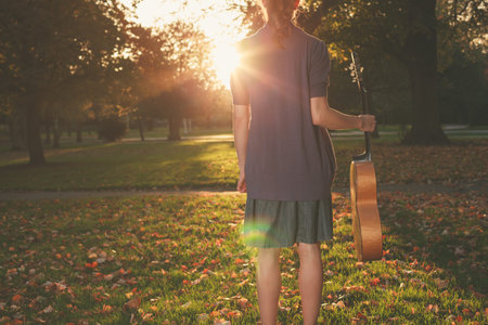Rear view shot of a young woman standing with a guitar in the park at sunset