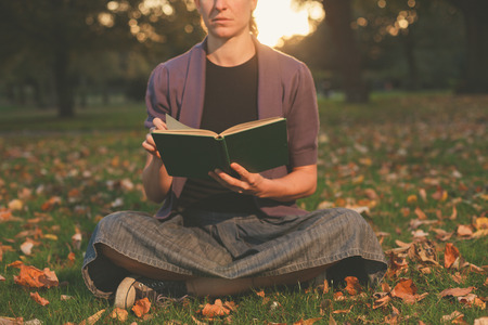 A young woman is sitting on the grass in the park and is reading a book at sunsetの写真素材
