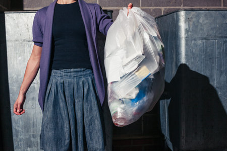 A young woman is standing next to some large bins with a bag of rubbish
