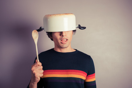 A silly young man is wearing a cooking pot on his had and is holding a wooden spoonの写真素材