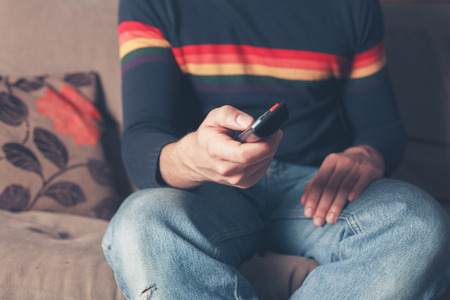 A young man is sitting on a sofa and is changing television channel with a remote controlの写真素材