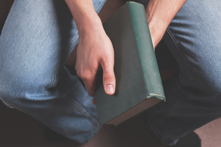 A young man is sitting and holding a big old bookの写真素材