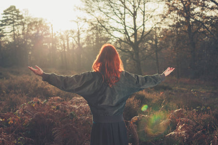 A young woman is raising her arms in joy as she is standing in a forest at sunsetの写真素材