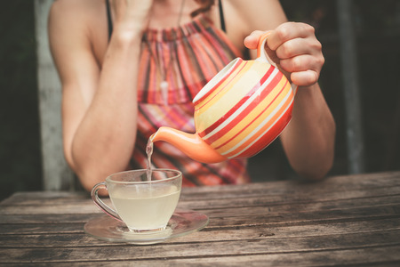 A young woman is sitting at a table and is pouring a cup of teaの写真素材