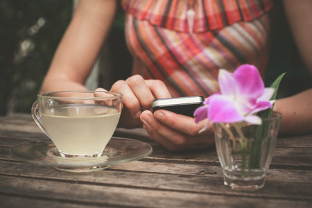 A young woman is drinking tea and using her smart phone at a wooden tableの写真素材