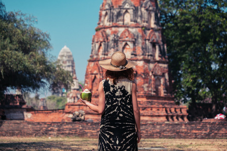 A young woman is drinking juice from a coconut near the ruins of an ancient buddhist templeの写真素材