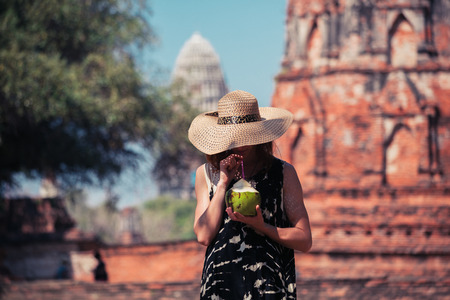 A young woman is drinking juice from a coconut near the ruins of an ancient buddhist templeの写真素材