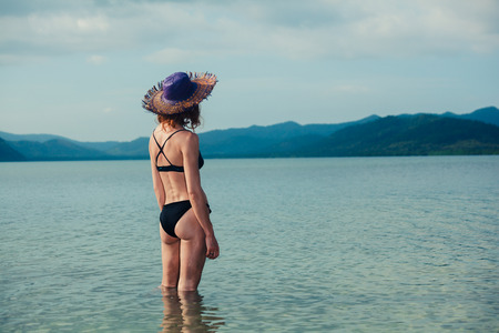 A young woman wearing a bikini is standing in the water and is looking at a tropical islandの写真素材