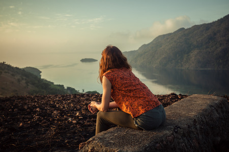 A young woman is sitting on an unusual rock on a mountain overlooking a bay at sunrise in a tropical climateの写真素材