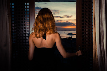 A sexy young woman wearing a dress is looking out at the sunset through her french doorsの写真素材