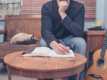 A young man is sitting with his cat and guitar at home on a sofa and is writing songs in a notebookの写真素材