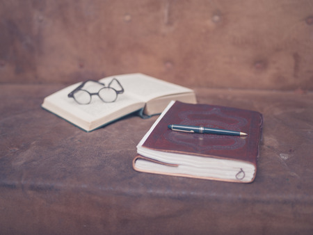 A pen on top of a notebook with an open book and a pair of glasses in the backgroundの写真素材