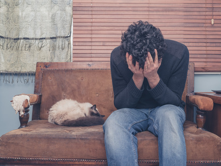 A sad and depressed young man is sitting on a sofa with a cat and his head buried in his handsの写真素材