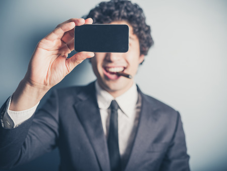 A young businessman is smoking a cigar and is taking a selfie using his smartphoneの写真素材