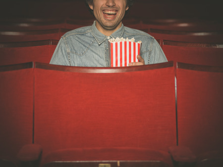 A happy young man with a bucket of popcorn is laughing and watching a movie at the cinemaの写真素材