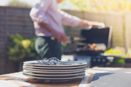 Stack of plates on a table outside in a garden with a man attending to a barbecue の写真素材