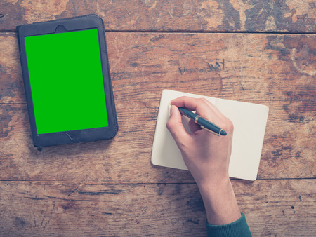 A male hand is writing in a notepad there is a tablet computer on the table as well with a green screenの写真素材