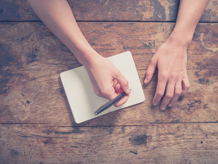 Close up on the hands of a young woman as she is writing in a small notepad at a wooden tableの写真素材