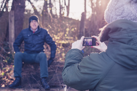 A young woman is taking a picture of a senior family member resting on a felled tree in a forest on a winter dayの写真素材
