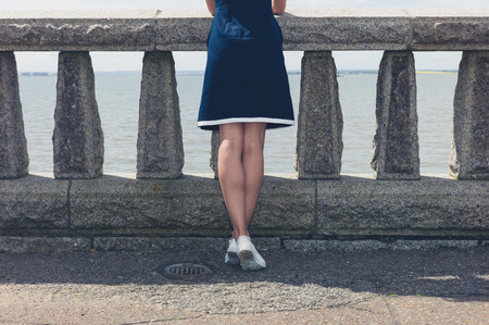 A young woman wearing a blue dress is standing by a wall with concrete balustrades on a promenade and is admiring the seaside on a sunny dayの写真素材