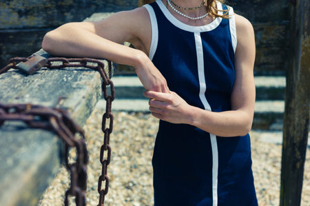 A young woman wearing a blue dress is standing on the beach on a sunny day by an old wooden...