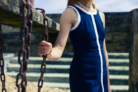 A young woman wearing a blue dress is standing on the beach on a sunny day by an old wooden structure with rusty chains hanging from itの写真素材