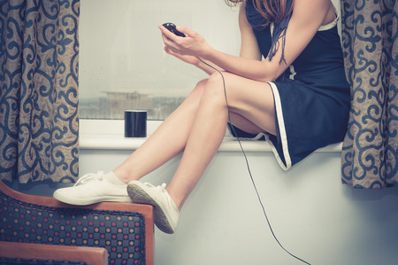 A young woman is sitting on a window sill and is charging her smartphoneの写真素材