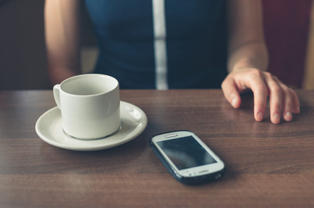 A young woman is sitting by the window in a diner with a cup and a smartphone in front of herの写真素材