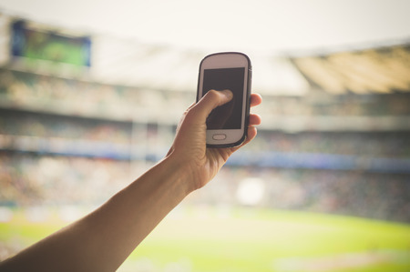 A female hand is holding a smart phone in a stadium to take pictures of a sporting eventの写真素材