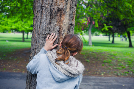 A young woman is hiding behind a tree in the parkの写真素材