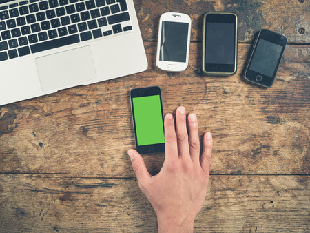 Overhead shot of a male hand using a smart phone with a green screen on a wooden table. There is a laptop and other smart phones on the table too.の写真素材