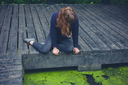 A young woman is sitting on a wooden deck by a dirty pond with algae in a forest