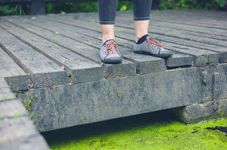 The feet of a young woman standing on a wooden deck by a dirty pond with algaeの写真素材