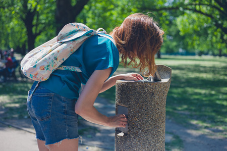 A young woman is drinking from a water fountain in a park on a sunny day in the summerの写真素材