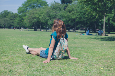 A young woman wearing a backpack is sitting on the grass and relaxing in a park on a summer dayの写真素材