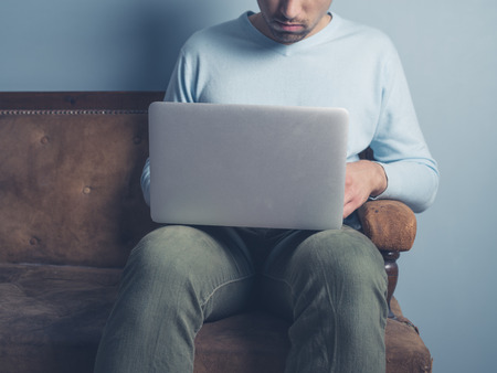 A young man is sitting on an old sofa and is using a laptop computerの写真素材