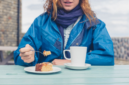 A young woman is having coffee and cake at a table outside on a cold dayの写真素材