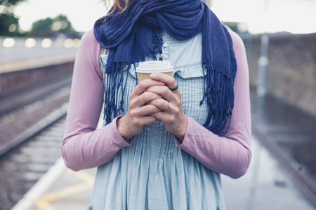 A young woman is standing on a platform with a cup of coffee and is waiting for the trainの写真素材