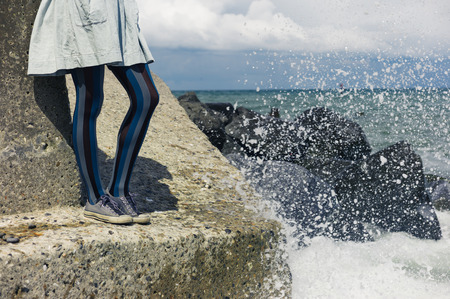 The legs of a young woman wearing a dress as she is standing on a rock by the coast with the waves crashing inの写真素材