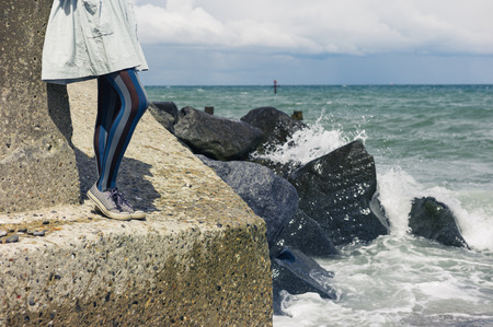 The legs of a young woman wearing a dress as she is standing on a rock by the coast with the waves crashing inの写真素材