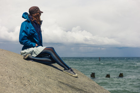 A young woman wearing a blue jacket is sitting on the flood defence by the sea