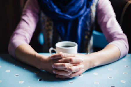 A young woman is sitting at a table with her hands folded around a cup of coffeeの写真素材