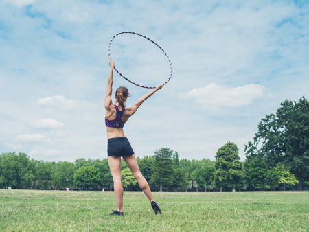 A fit and athletic young woman is standing on the grass in a park with a hoop raised above her headの写真素材