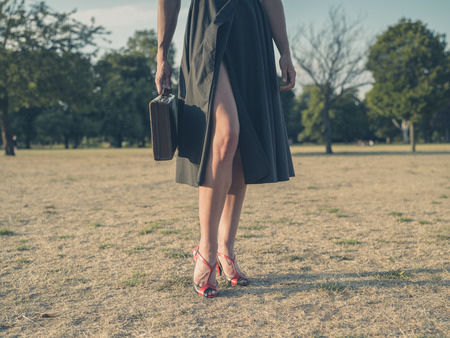 Vintage filtered shot of an elegant young woman wearing a dress and high heels standing in a park with a briefcaseの写真素材