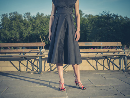 Vintage filtered shot of an elegant young woman wearing a dress and standing by some park benchesの写真素材