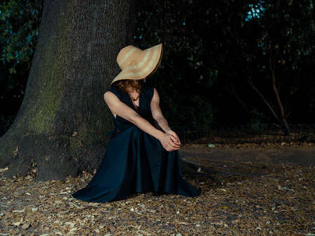 An elegant woman wearing a dress and a hat is relaxing under a tree in the parkの写真素材