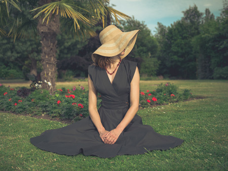 Vintage filtered shot of an elegant woman with a hat sitting on the grass in a park by a flowerbedの写真素材